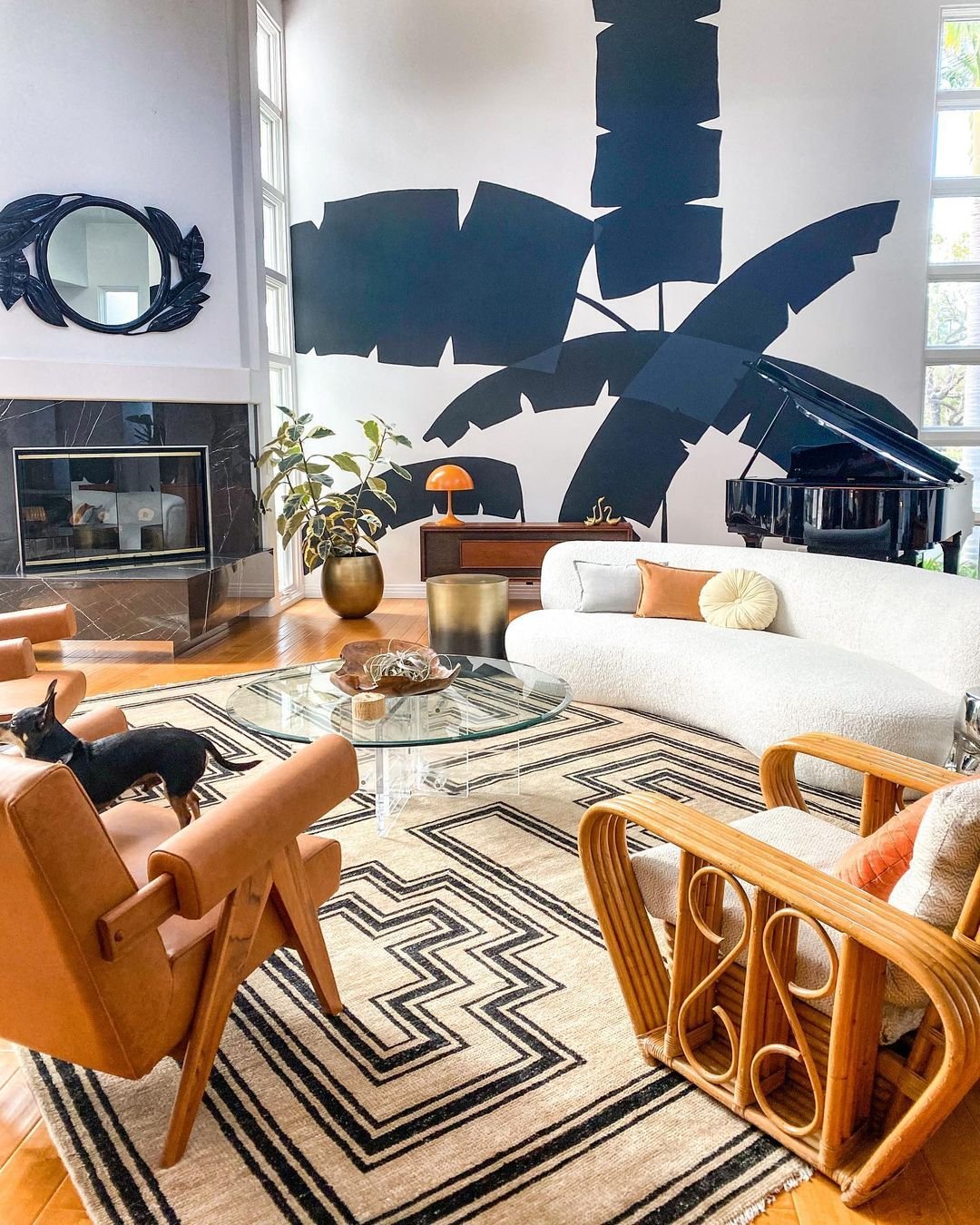 A low ivory boucle curved sofa sits atop the natural and black geometric Senna rug in this modern living room. Two leather arm chairs and a rattan chair sit across from the sofa, and a glass coffee table is in the middle A large grand piano, writing desk, and a large blue palm leaf mural are behind the sofa.