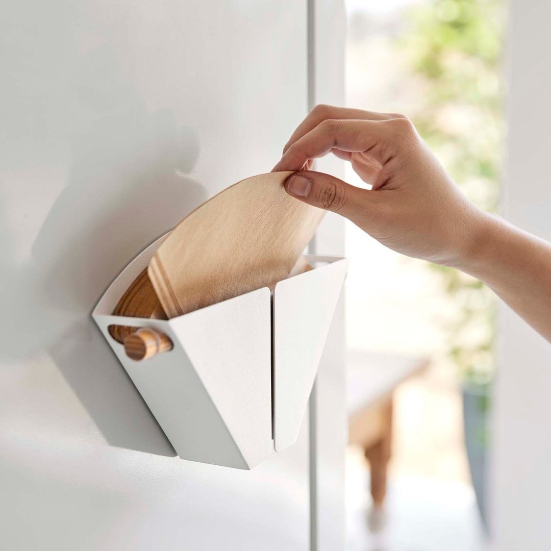 A hand reaching for a coffee filter from a white magnetic coffee filter by Yamazaki Home on the side of a fridge.