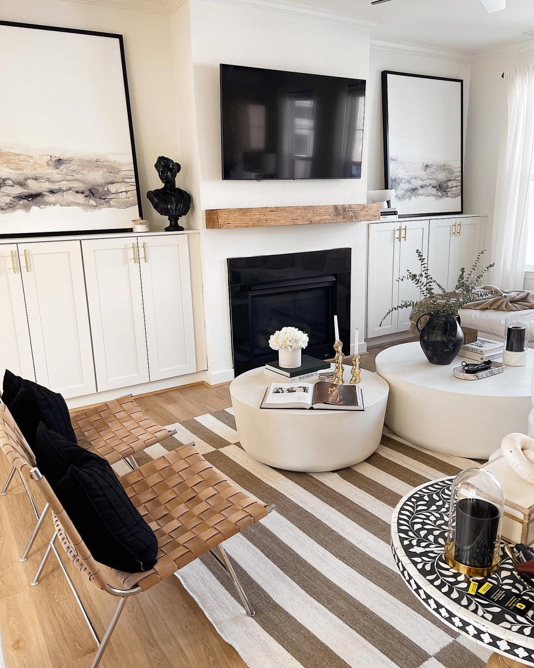 Two woven leather armless chairs sit atop an offset brown and white striped rug. Two round white coffee tables hold vases, books and candles and sit in front of the chairs, while the wall next to the chairs has a fireplace and built-in white cabinets  and large black and white abstract art.