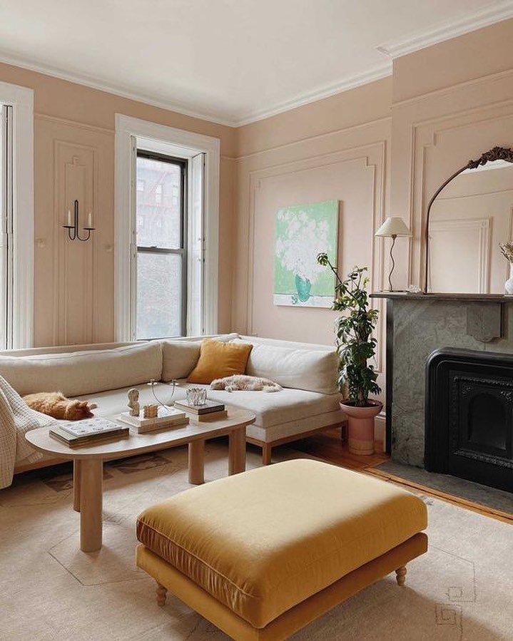 Blush walls with paneling give this living room a classif feel. The low ivory linen sofa on a wooden base and a light wood coffee table modernize the room. A gold upholstered ottoman sits across from a black stone fireplace.