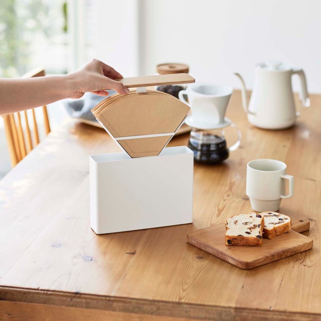 Coffee Filter Case by Yamazaki Home in white on a dining table. A hand is lifting the top to reveal coffee filters inside.