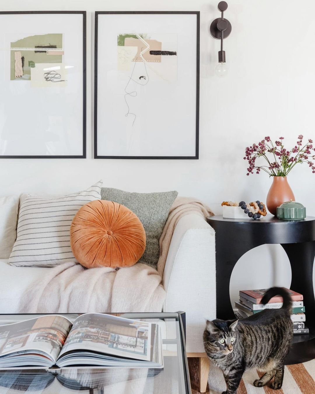 Two large framed abstract art pieces hang above an ivory sofa with a gray, white and gray striped, and round orange throw pillows. A throw blanket is draped over the sofa. Next to the sofa is a round black end table with an orange vase with small flowers and small decorative objects. A tabby cat walks in front.