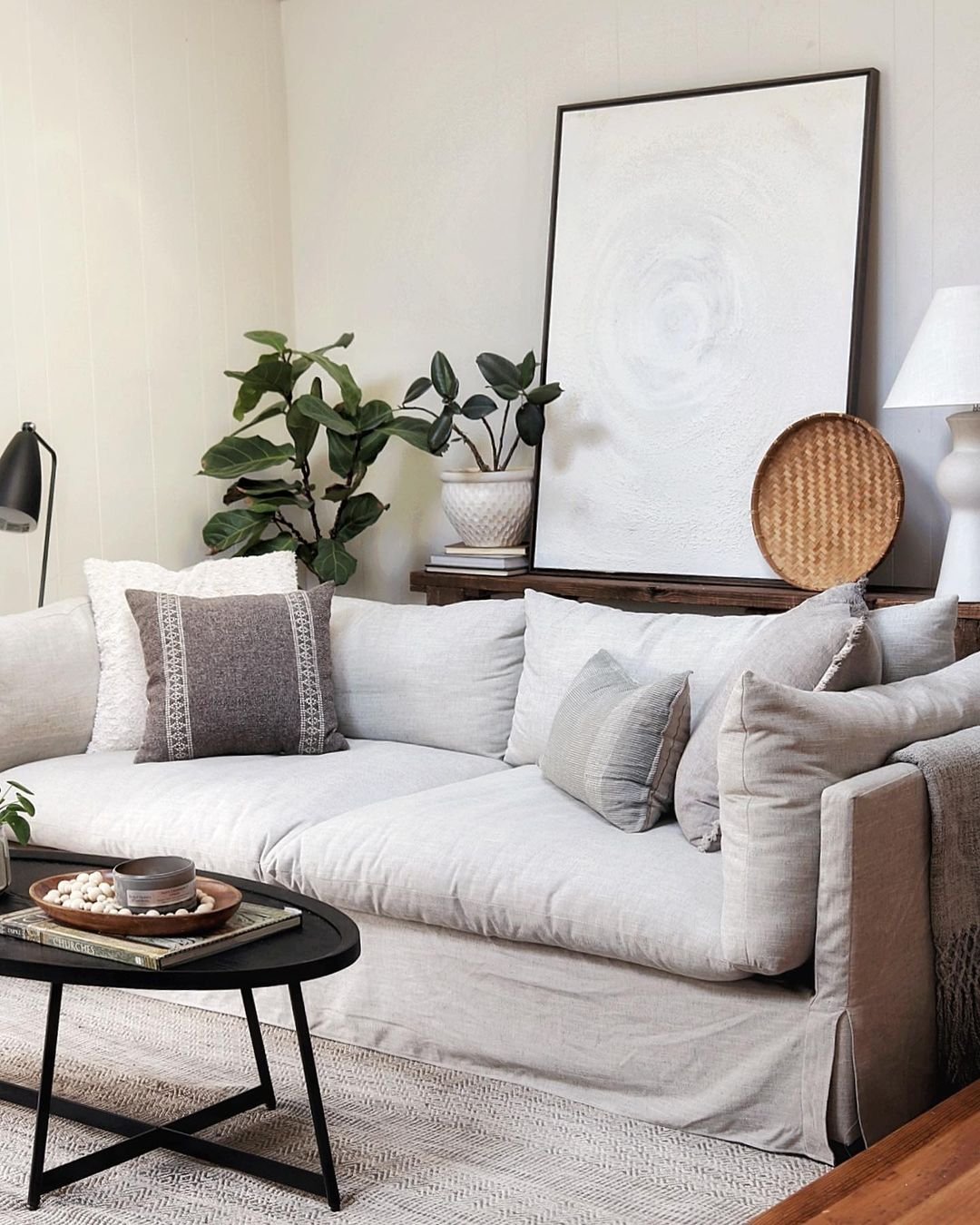 A linen slipcovered sofa with neutral throw pillows has a wooden console table behind it holding a tall white geometric painting, a plant, woven basket and a white lamp. A dark wood coffee table sits in front of the sofa.