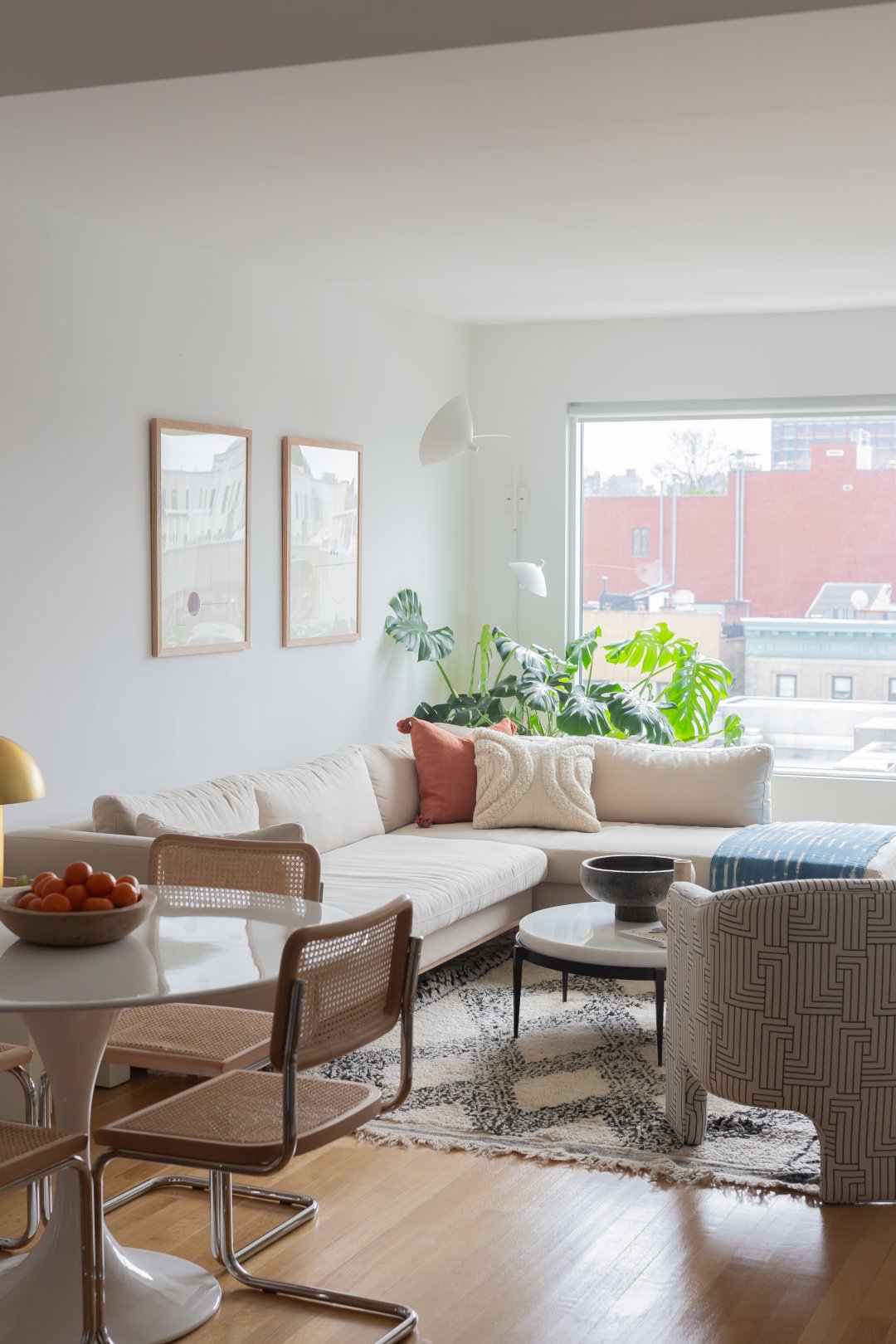 A low ivory sofa with chaise has a blue patterned throw blanket, white throw pillow and orange throw pillow. A rounded stone topped coffee table with a black base sits in the middle and a gemetric white and black patterned arm chair sits opposie the sofa. A round white table surrounded by cane chairs sits next to the sofa.