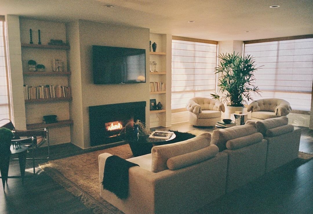 A light colored sofa sits in front of a fireplace wall with a tv hung above it. Two built-in bookcases flank the fireplace. Two rounded tufted accent chairs sit off to the side with a large plant in between them.