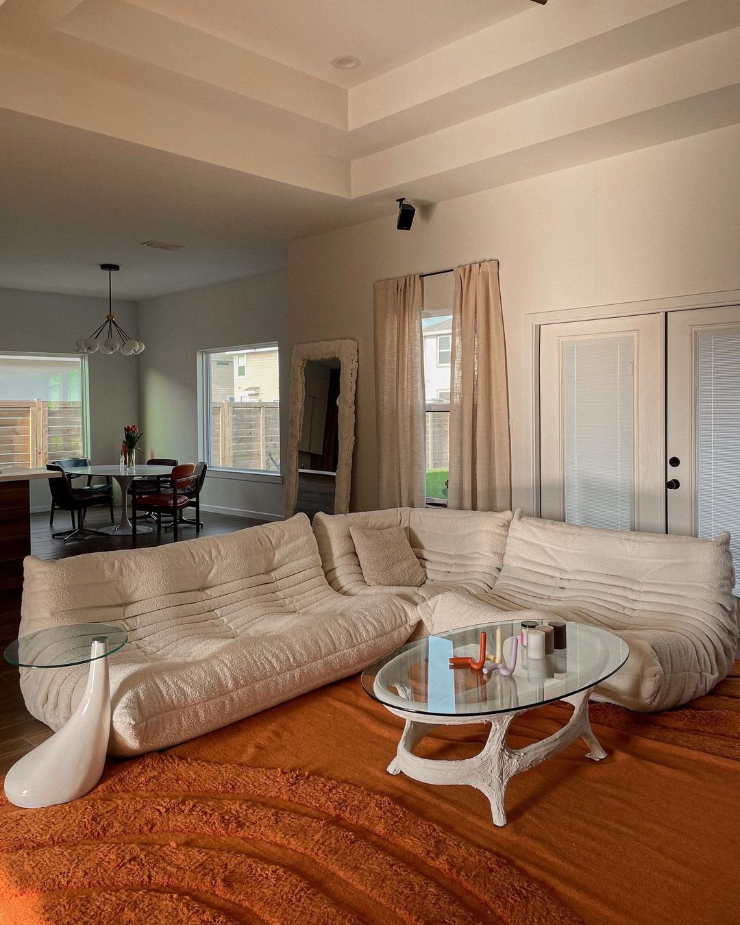 A low armless tan boucle sectional sofa sits atop a rust-colored Arches rug. A glass coffee table with molded bottom sits in the middle and holds different candles.